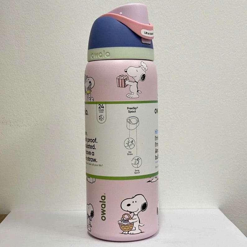 May include: A pink Owala FreeSip water bottle featuring Snoopy illustrations. The bottle has a blue and pink lid with a carrying handle. It is insulated and has a straw. The bottle is designed to keep drinks cold for 24 hours.