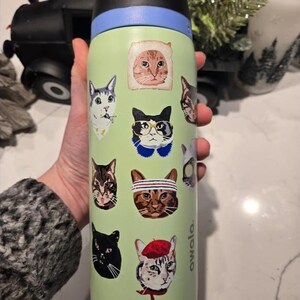 May include: A light green Owala water bottle featuring various cat stickers. The designs include a cat in a bread slice, a cat wearing glasses, a cat with a headband, a cat in a red beret, and several other feline portraits in different styles.