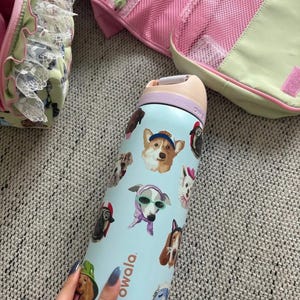 Pu&ograve; includere: Una borraccia Owala azzurra con un motivo ripetuto di varie razze canine che indossano accessori come cappelli, occhiali da sole e sciarpe. La bottiglia ha un tappo color pesca con un anello viola e una maniglia per il trasporto.