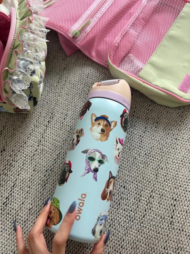 Pu&ograve; includere: Una borraccia Owala azzurra con un motivo ripetuto di varie razze canine che indossano accessori come cappelli, occhiali da sole e sciarpe. La bottiglia ha un tappo color pesca con un anello viola e una maniglia per il trasporto.