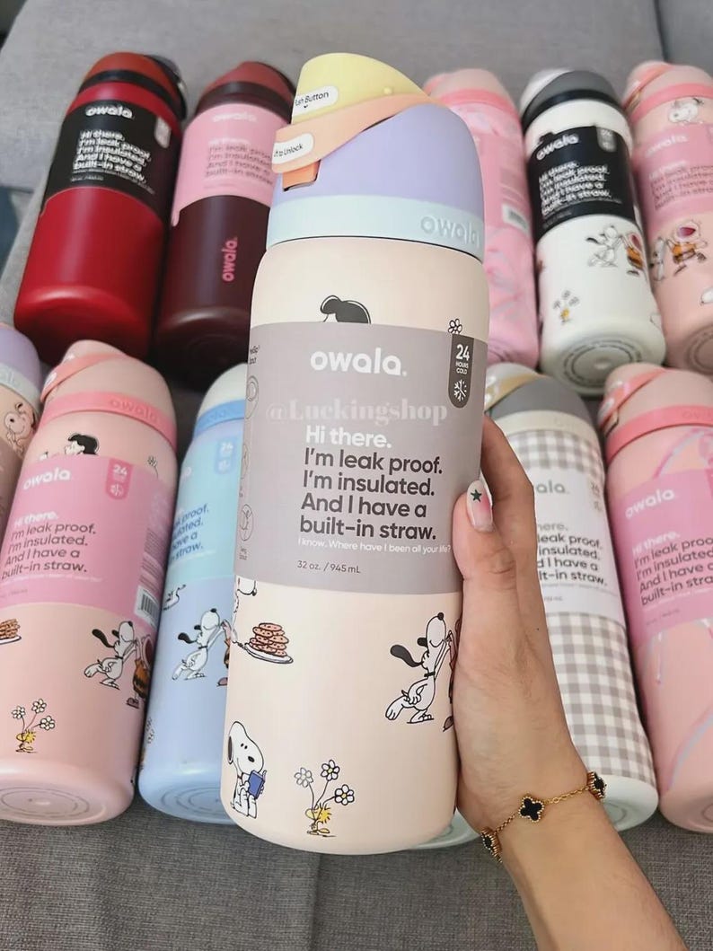 May include: A collection of Owala insulated water bottles in various colors, including pink, red, and white. The bottles feature a built-in straw and Snoopy graphics. One bottle is held by a hand, showcasing the text "Hi there. I'm leak proof. I'm insulated."