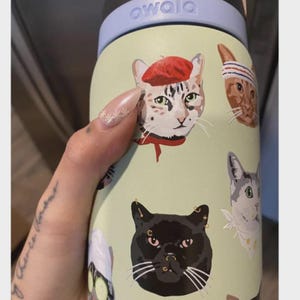May include: A light green Owala water bottle featuring a repeating pattern of various cat illustrations. The cats are depicted with different accessories, including a red beret, a striped headband, and facial piercings on a black cat.