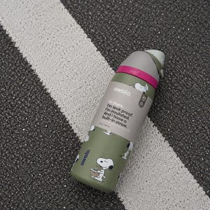 Puede incluir: Una botella de agua Owala verde oliva con un dise&ntilde;o de dibujos animados de Snoopy. La botella tiene una tapa gris y una banda rosa. El texto en la botella dice: "Hola. Soy a prueba de fugas. Estoy aislado. Y tengo una pajita incorporada." Capacidad: 946 ml.