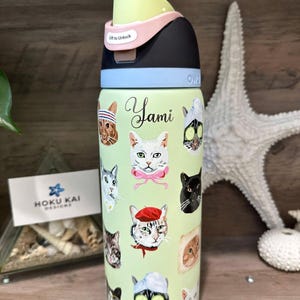 May include: A light green stainless steel water bottle featuring a variety of cat illustrations, including cats wearing berets, towels, and cucumber slices. The name Yami is printed in black script. The lid is black, light blue, and pale yellow.