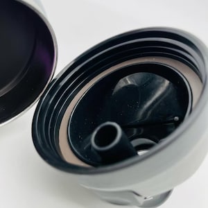 May include: Close-up of a black and silver insulated tumbler lid. The lid is open, revealing a black interior with a drinking spout. A brown gasket seals the lid. The tumbler's exterior is silver.