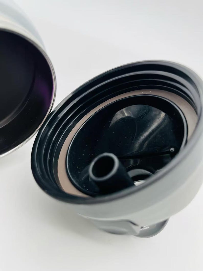May include: Close-up of a black and silver insulated tumbler lid. The lid is open, revealing a black interior with a drinking spout. A brown gasket seals the lid. The tumbler's exterior is silver.