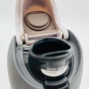 May include: Close-up of a gray and white water bottle with a flip-top lid. The lid is open, revealing the drinking spout and interior. The bottle's design includes a gray body and a black spout.