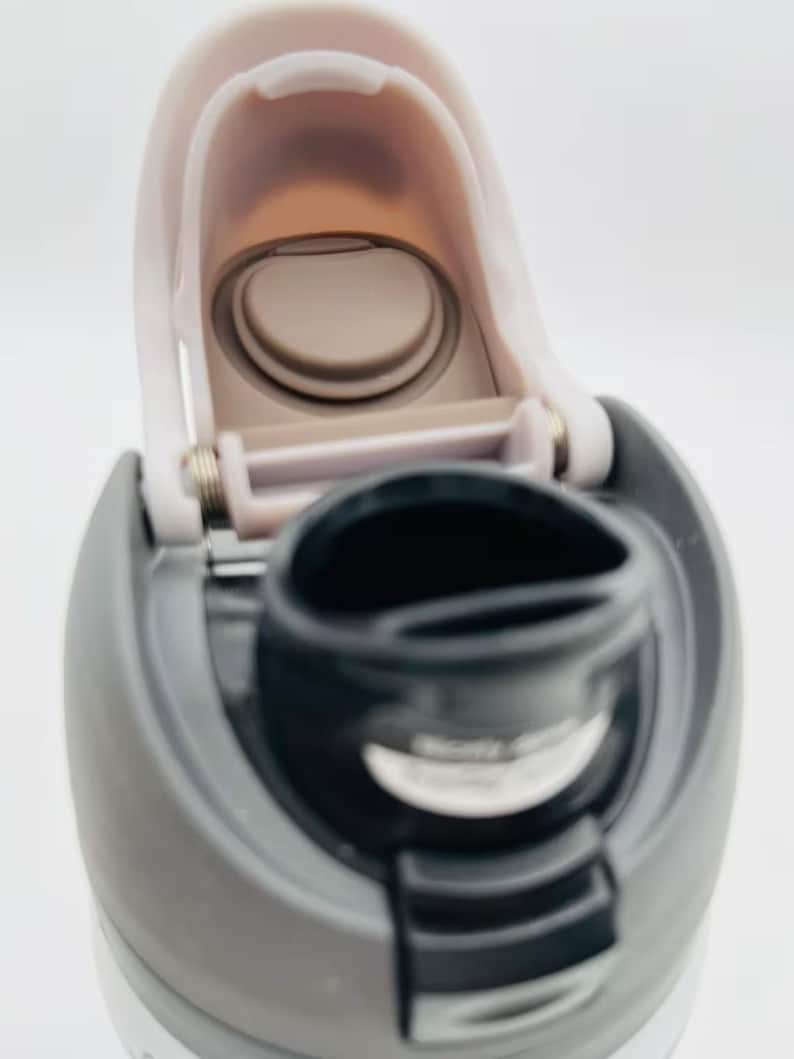 May include: Close-up of a gray and white water bottle with a flip-top lid. The lid is open, revealing the drinking spout and interior. The bottle's design includes a gray body and a black spout.