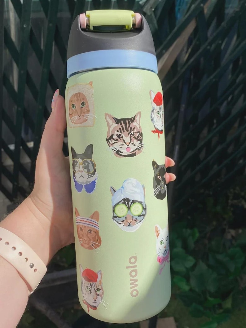 May include: A light green Owala water bottle featuring various cat illustrations, including a cat in a bread slice, a cat with a towel and cucumber slices on its eyes, and cats wearing hats. The bottle has a black and light blue lid with a yellow latch.