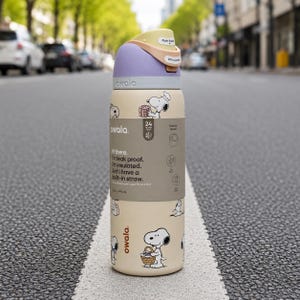 Puede incluir: Una botella de agua Owala beige con tapa morada y gr&aacute;ficos de dibujos animados de Snoopy. La botella tiene una pajita incorporada y es a prueba de fugas y aislada. La botella tiene el texto "Hola. Soy a prueba de fugas. Estoy aislado. Y tengo una pajita incorporada."
