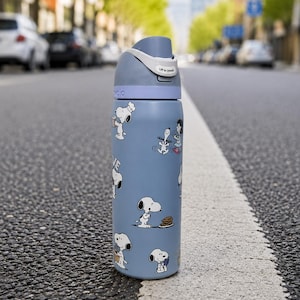 Puede incluir: Una botella de agua azul con personajes de Snoopy. La botella tiene una tapa gris con un asa blanca y el texto "Lift to Unlock". La botella est&aacute; sobre una carretera.