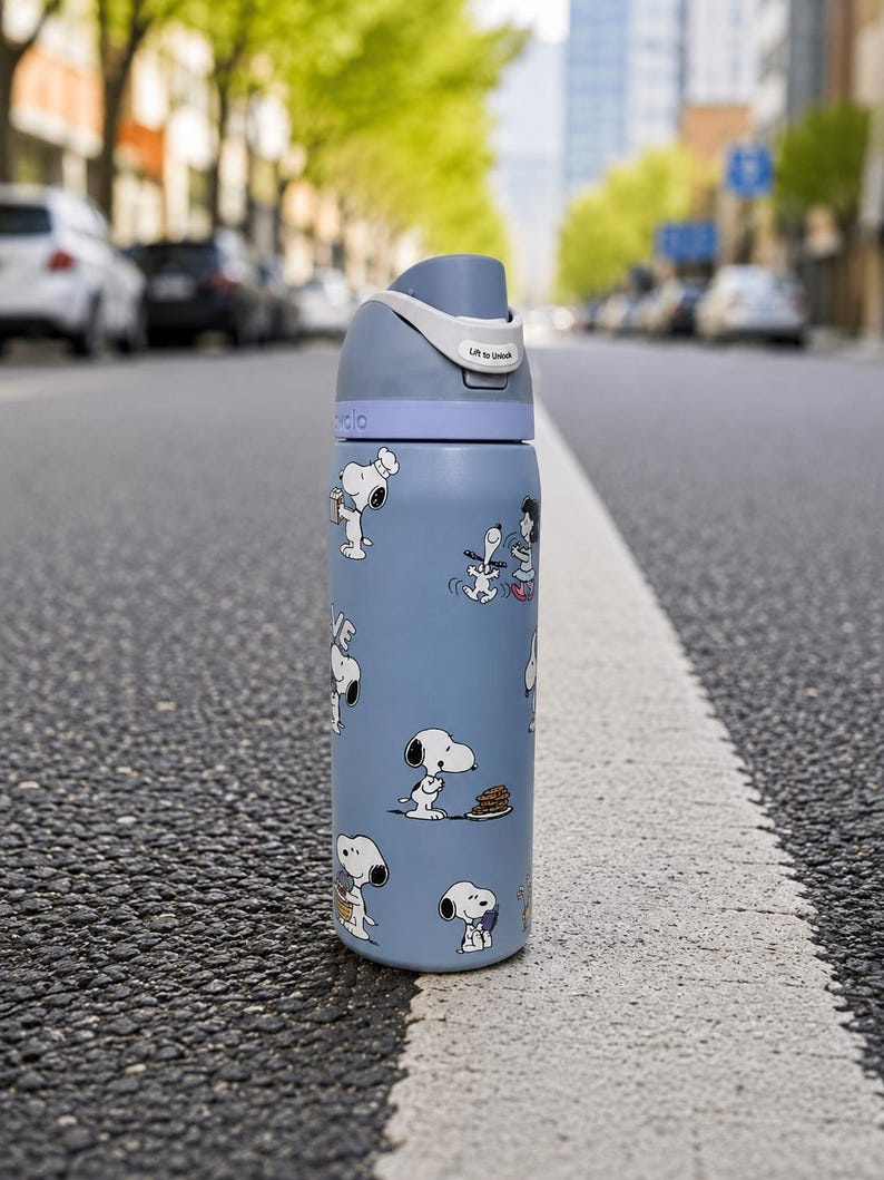 Puede incluir: Una botella de agua azul con personajes de Snoopy. La botella tiene una tapa gris con un asa blanca y el texto "Lift to Unlock". La botella est&aacute; sobre una carretera.