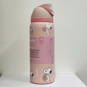 May include: A pink Owala water bottle featuring Snoopy illustrations. The bottle has a flip-top lid and a built-in straw. The bottle is leak-proof and insulated, with a capacity to keep drinks cold for 24 hours.