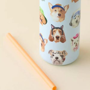 Pu&ograve; includere: Un bicchiere cilindrico azzurro con un motivo ripetitivo di varie razze canine che indossano cappelli e accessori, tra cui un cane con la scritta Good Boy 777. Una cannuccia arancione &egrave; posizionata in diagonale accanto al bicchiere.