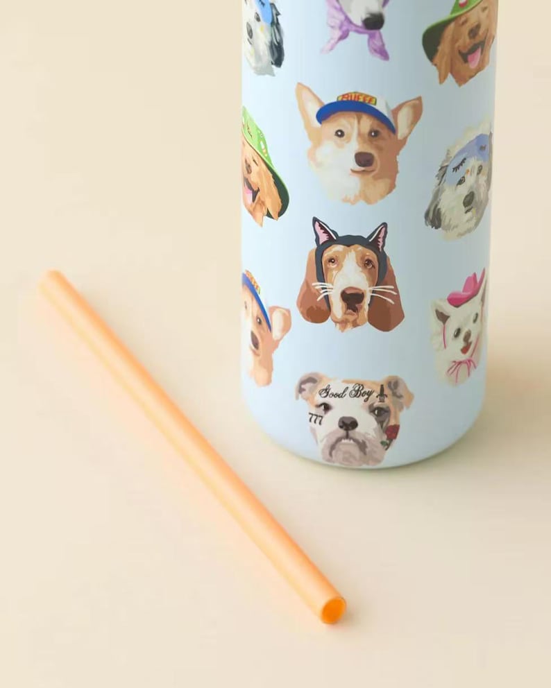 Pu&ograve; includere: Un bicchiere cilindrico azzurro con un motivo ripetitivo di varie razze canine che indossano cappelli e accessori, tra cui un cane con la scritta Good Boy 777. Una cannuccia arancione &egrave; posizionata in diagonale accanto al bicchiere.