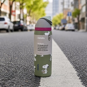 Puede incluir: Una botella de agua Owala verde y gris con un dise&ntilde;o de Snoopy y el texto "Hi there. I'm leak proof. I'm insulated. And I have a built-in straw." La botella es de 295 ml.