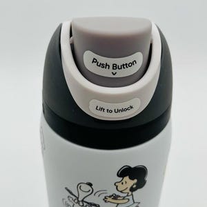 May include: White and black insulated water bottle featuring a cartoon design of Snoopy and Lucy. The lid has a gray push button and the text "Push Button" and "Lift to Unlock".