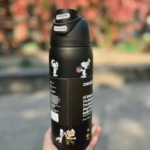 May include: Black Owala water bottle featuring Snoopy and Woodstock graphics. The bottle has a push-button lid and text that reads "Hi there, I'm lead, I'm in, And I'm built-". The bottle holds 32 oz.