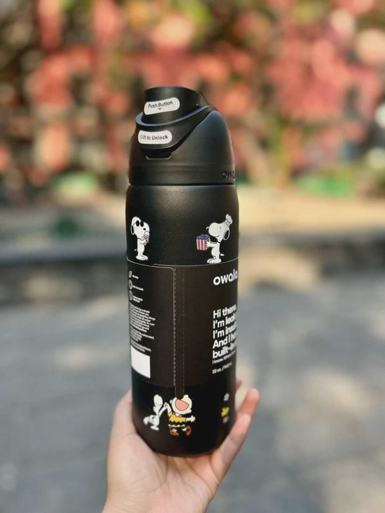 May include: Black Owala water bottle featuring Snoopy and Woodstock graphics. The bottle has a push-button lid and text that reads "Hi there, I'm lead, I'm in, And I'm built-". The bottle holds 32 oz.