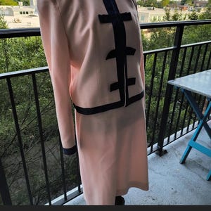 May include: A pink and black women's suit with a skirt and jacket. The jacket has a black trim and a black bow on the front. The skirt is a-line and has a black trim at the hem.