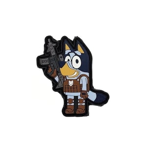 May include: A rubberized patch featuring a cartoon blue dog character wearing a brown tactical vest and holding a black rifle. The character has light blue fur, dark blue ears, and a yellow snout, rendered in a flat, graphic style with black outlines.