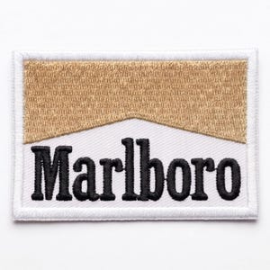 May include: A rectangular embroidered patch featuring a gold-colored upper section and a white lower section. The word Marlboro is stitched in bold black lettering across the white portion. The entire patch is bordered by a white stitched edge.