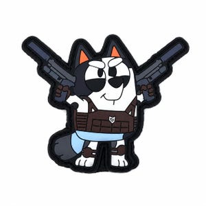 Puede incluir: Un parche de goma que muestra un personaje de perro de dibujos animados con pelaje blanco y negro, puntas de orejas naranjas y una cola azul claro. El personaje lleva un chaleco táctico marrón y sostiene dos pistolas gris oscuro.