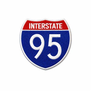 May include: A shield-shaped embroidered patch featuring the text INTERSTATE in white on a red background at the top, with the number 95 in large white digits on a blue background below, all bordered by a white stitched edge.