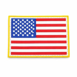 Puede incluir: Un parche rectangular que muestra la bandera de los Estados Unidos con un cantón azul que contiene estrellas blancas y franjas horizontales rojas y blancas. El diseño está bordeado por una línea blanca fina y un marco exterior amarillo grueso.