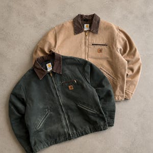May include: Two Carhartt zip-up jackets, one in tan and one in dark green, both featuring brown corduroy collars, front zipper closures, two lower hand pockets, and a small zippered chest pocket with a square Carhartt brand logo patch.