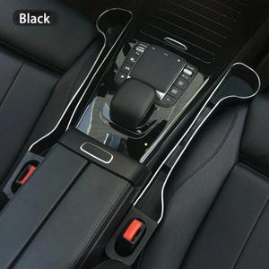 May include: Black car interior showing a centre console, armrest, and seat belts. The console has a control panel and a glossy black finish. Silver trim accents the console and seat dividers. The word "Black" is in the upper left corner.