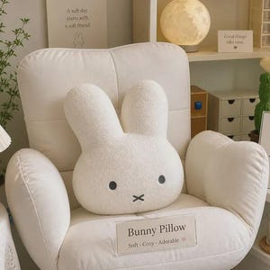 May include: A white plush pillow shaped like a rabbit head with two tall ears, two small black circular eyes, and a black cross-shaped mouth. The pillow is positioned on a white upholstered chair and features a label reading Bunny Pillow.