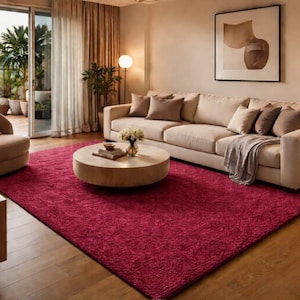 May include: A large, rectangular, deep magenta area rug with a textured, shaggy pile surface. The rug is positioned on a light-colored wooden floor in a living room, centered beneath a round wooden coffee table and a long, beige upholstered sofa.