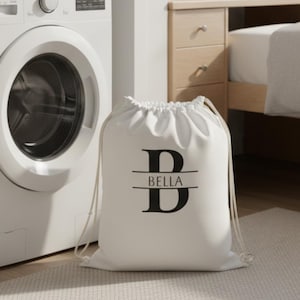 May include: A white drawstring laundry bag featuring a black monogram design with the letter B and the name BELLA printed in the center. The bag is positioned on the floor next to a stacked white washer and dryer unit in a room setting.