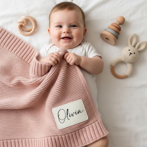 May include: A dusty pink knitted baby blanket with a ribbed border and a rectangular white patch featuring the name Olivia in black script. The blanket is accompanied by a wooden ring toy, a wooden stacking toy, and a crocheted bunny rattle.