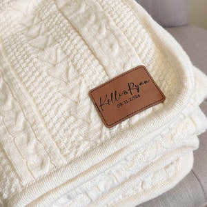 May include: Cream-colored cable knit blanket with a personalized leather patch. The patch is brown and features the names "Kelli & Ryan" and the date "08.11.2024". The blanket is folded and resting on a gray surface.