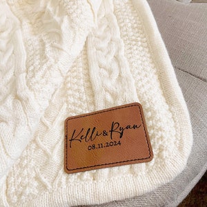 May include: A white cable knit throw blanket with a brown leather patch that says "Kelli & Ryan 08.11.2024"