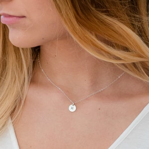 Op de afbeelding: Een delicate zilveren ketting met een kleine, ronde hanger met een gegraveerde initiaal. De hanger hangt aan een fijne ketting, wat een minimalistische en elegante uitstraling cre&euml;ert. De ketting wordt gedragen met een witte top.
