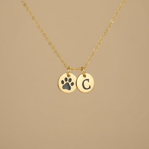 Op de afbeelding: Een goudkleurige ketting met twee ronde hangers. E&eacute;n hanger heeft een zwarte pootafdruk en de andere de letter "C" in zwart. De ketting is ook goudkleurig.