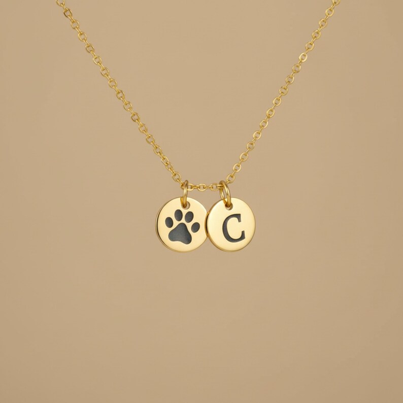 Op de afbeelding: Een goudkleurige ketting met twee ronde hangers. E&eacute;n hanger heeft een zwarte pootafdruk en de andere de letter "C" in zwart. De ketting is ook goudkleurig.