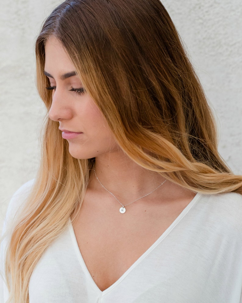 Op de afbeelding: Een delicate zilveren ketting met een kleine, ronde hanger met een gegraveerde initiaal. De hanger hangt aan een fijne ketting, wat een minimalistische en elegante uitstraling cre&euml;ert. De ketting wordt gedragen met een witte top.