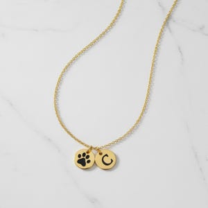 Op de afbeelding: Een goudkleurige ketting met twee ronde hangers. E&eacute;n hanger heeft een zwarte pootafdruk en de andere de zwarte letter "C". De ketting wordt gepresenteerd op een wit marmeren oppervlak.
