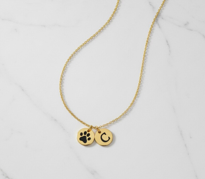 Op de afbeelding: Een goudkleurige ketting met twee ronde hangers. E&eacute;n hanger heeft een zwarte pootafdruk en de andere de zwarte letter "C". De ketting wordt gepresenteerd op een wit marmeren oppervlak.