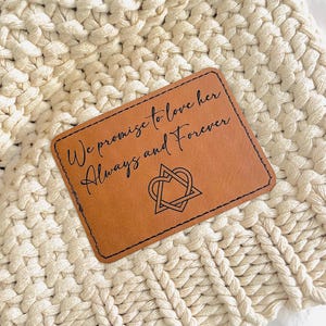 May include: A rectangular, brown leather patch with the words "We promise to love her Always and Forever" in black script. A heart-shaped design is below the text. The patch is set against a cream-colored, woven background.