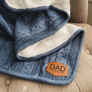 May include: A blue knitted throw blanket with a white sherpa lining. The blanket has a brown leather patch with the word "DAD" and the names "ryan.alexis.max" in smaller font.
