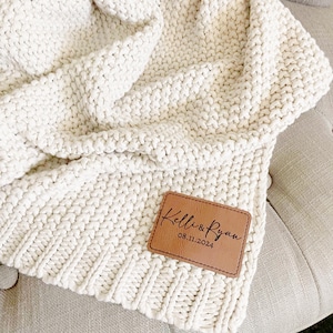 May include: Cream-colored, chunky knit blanket with a personalized leather patch. The patch is brown and features the names "Kelli & Ryan" and the date "08.11.2024" in elegant script. The blanket is draped over a neutral-toned surface.