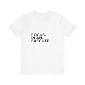 Może przedstawiać: Biały t-shirt z krótkim rękawem z czarnym napisem FOCUS. PLAN. EXECUTE. nadrukowanym czystą czcionką bezszeryfową na środku klatki piersiowej. Odzież posiada klasyczny okrągły dekolt oraz standardowy krój, zapewniając prosty wygląd.