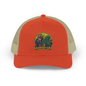 May include: A trucker hat with a red front panel and a tan mesh back. The hat features a colorful graphic of a diver and an octopus with the text "SEA BRETHREN" on a banner.