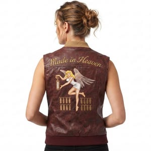 May include: A sleeveless brown faux leather vest featuring a tan ribbed collar and hem. The back displays an embroidered graphic of a blonde angel holding a bomb, with the text Made in Heaven above and rows of small bomb icons below the figure.