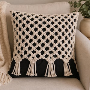 Pode incluir: Uma almofada quadrada com uma base de tecido preto sobreposta por um intrincado padrão de corda de macramé na cor creme. O design de macramé cria uma estrutura de treliça de diamantes repetitivos que termina na borda inferior com uma série de borlas de franjas decorativas.